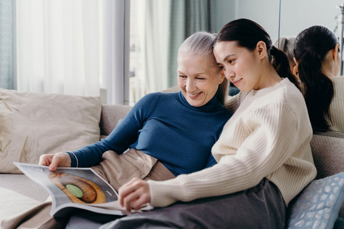 Two women sitting on a sofa, smiling and looking at a book, illustrating parents and daughter relationship moments.