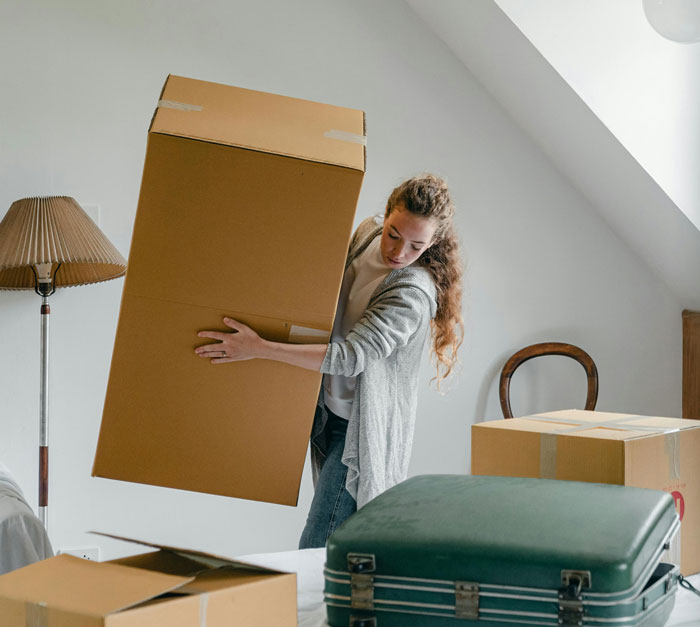 Young woman carrying a large moving box amid packed suitcases and boxes in a remodeled room during a move out.
