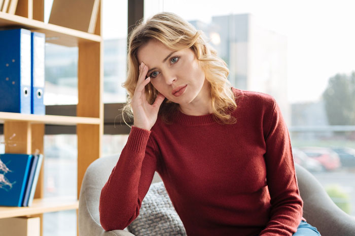 Woman in a red sweater looking worried and thoughtful, sitting indoors near a window in a bright room.