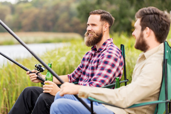 Two men fishing by the lake on a boys trip, one holding a fishing rod and a beer bottle, enjoying outdoors.