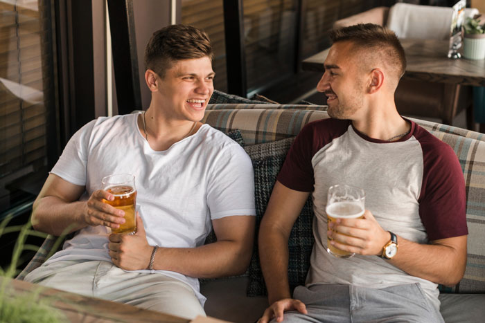 Two men enjoying beers and laughing together on a couch during a casual boys trip outing.