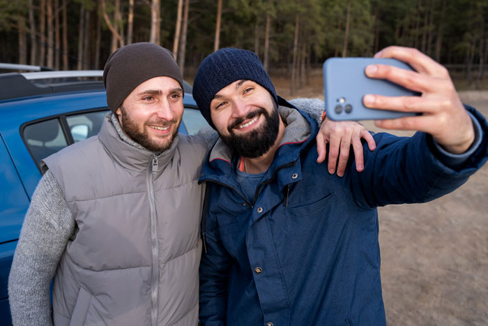 Two men smiling and taking a selfie outdoors during a boys trip in a forested area near a car.