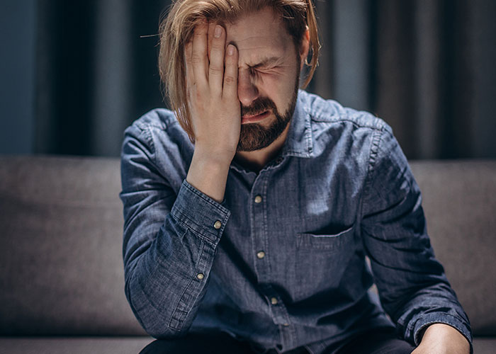 Man in denim shirt sitting on couch with hand on face, expressing distress and villain emotions in storytelling context.