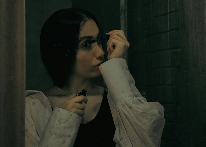 Young woman applying mascara in a dimly lit room, reflecting on the villain in someone else’s story theme.