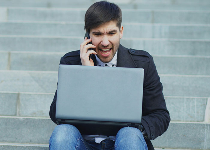 Young man looking angry while talking on phone and using laptop sitting on outdoor stairs portraying villain behavior online