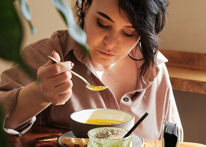 Woman eating soup at wooden table, reflecting quietly, capturing the mood of a villain in someone else’s story.