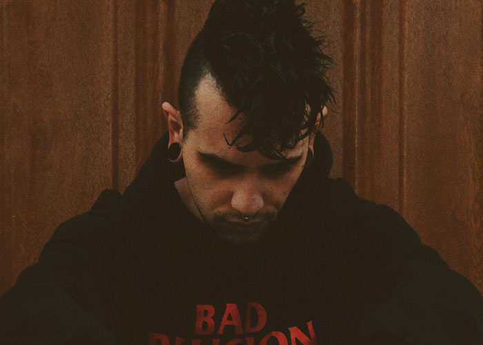 Young man with mohawk hairstyle and stretched ears sitting against a wooden background, embodying a villain persona.