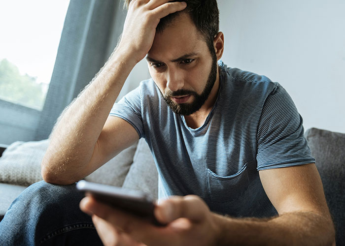 Man with worried expression sitting on couch, holding phone, reflecting on being the villain in someone else's story.