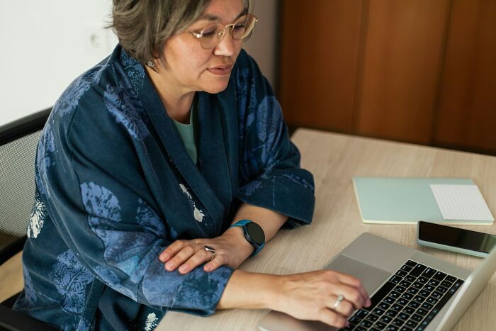 Person in a blue patterned jacket and glasses sharing chaotic breakup stories while working on a laptop at a wooden desk.