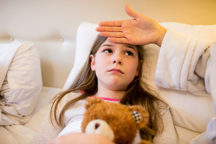 Young girl lying on a couch holding a teddy bear while an exhausted mom checks her forehead for a fever.