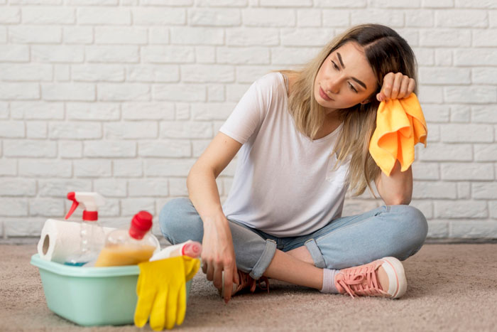 Exhausted mom sitting on floor with cleaning supplies, holding a cloth, overwhelmed by house chores and parenting.