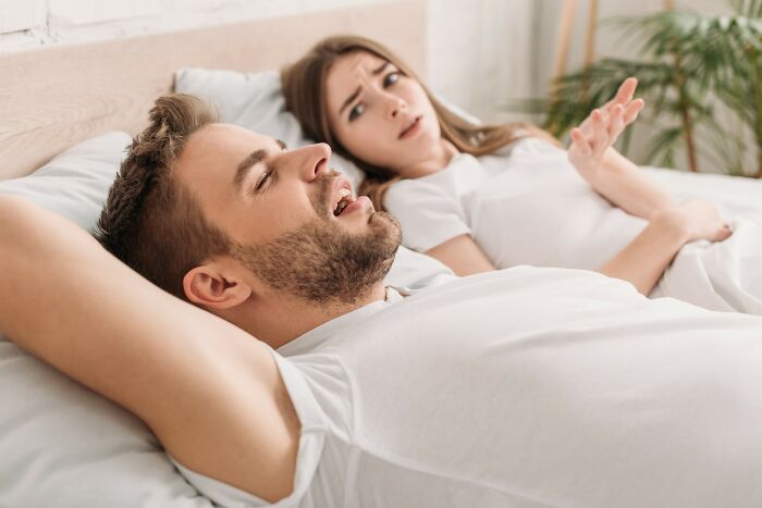 Man lying in bed snoring loudly while woman looks frustrated illustrating medical problems far worse than people think.