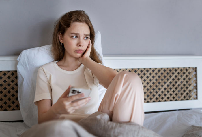 Woman obsessed with being a bonus mom feeling crushed sitting on bed holding a phone with a worried expression.