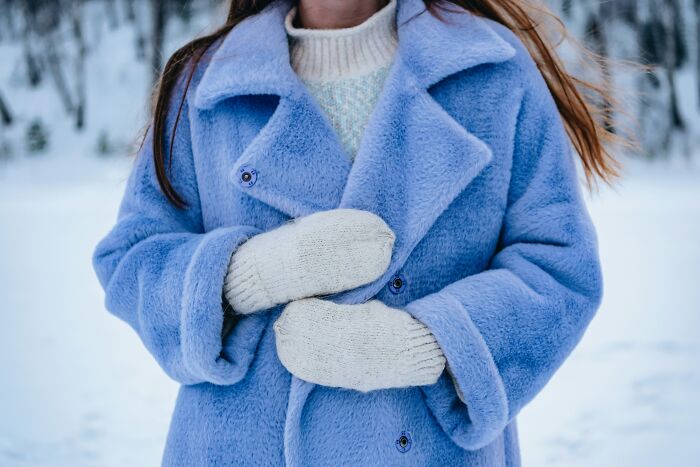 Person wearing a warm blue coat and white mittens standing outdoors in a snowy landscape during winter.