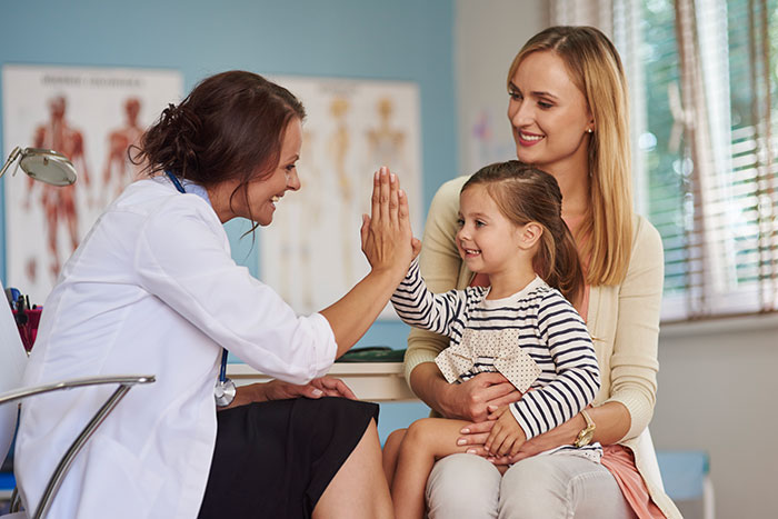 Pediatrician giving high five to a young girl while mother looks on during a child&rsquo;s medical procedure visit.