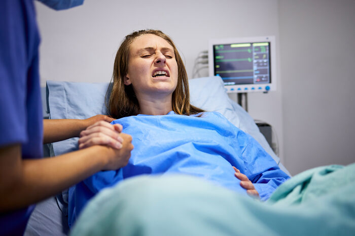 A woman in labor experiencing pain during birth while a medical professional holds her hand in a hospital room.