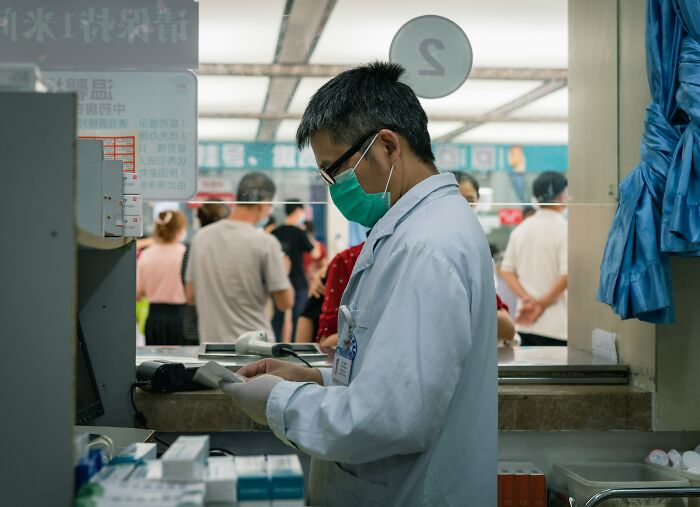 Hospital worker in a mask and white coat handling documents inside a busy hospital with patients in the background.
