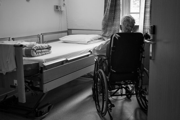 Elderly person in a wheelchair looking out hospital window near an empty bed, reflecting hospital workers' creepiest experiences.