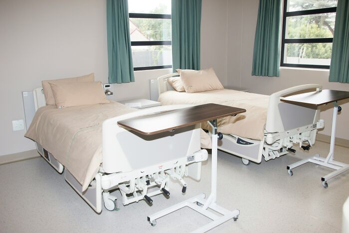 Two empty hospital beds with beige linens and bedside tables in a quiet hospital room during the day