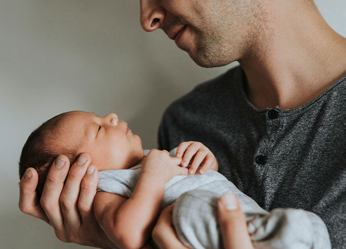 Hombre sosteniendo un recién nacido en brazos en una habitación de parto, momento cargado de emociones y caos después del engaño.