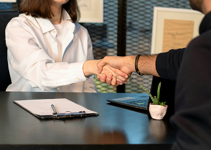 Two people shaking hands across a table with a clipboard and laptop, symbolizing benefits for women to get married.