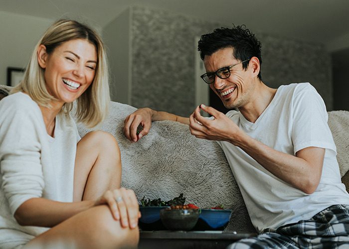 Couple laughing together on couch, discussing real benefits for women to get married in a relaxed home setting.