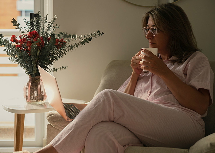 Woman in pink pajamas sipping coffee while looking at laptop, reflecting on benefits of marriage for women.