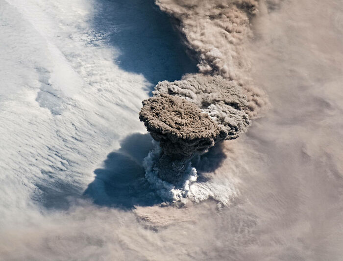 Volcanic ash plume rising amid clouds, showcasing the beauty and mystery of space and earth from above.