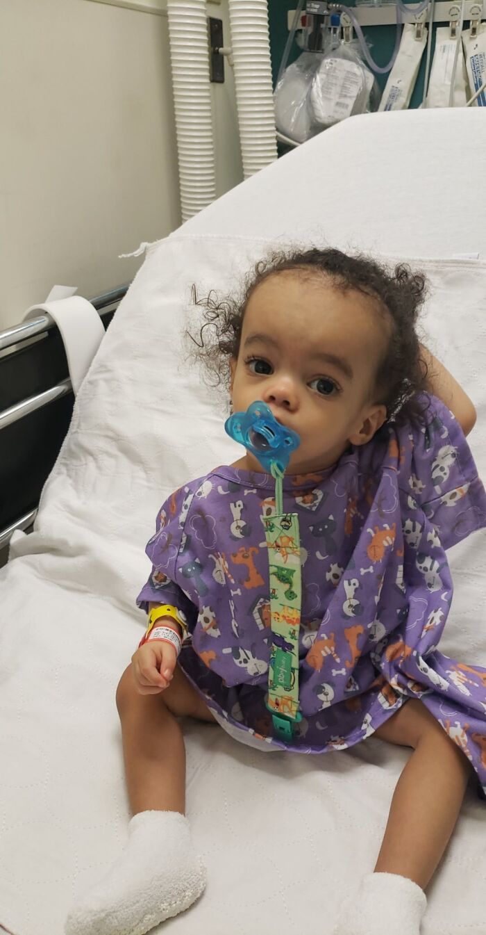 Young child in hospital gown with pacifier, sitting on medical bed surrounded by medical equipment in clinical setting.