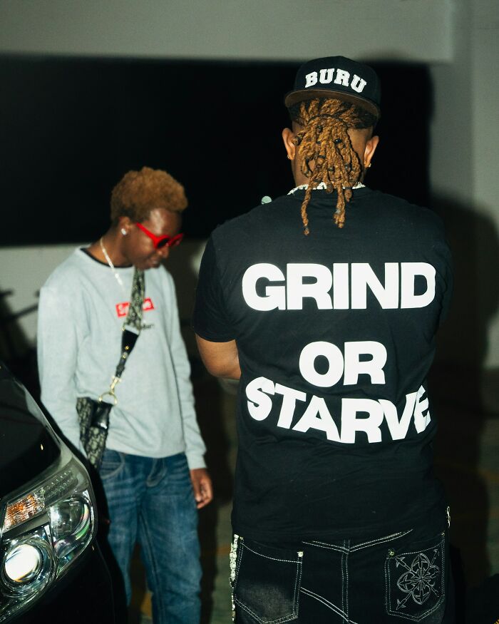 Two men standing near a car, one wearing a shirt with grind or starve text, reflecting healthy habits discussion.