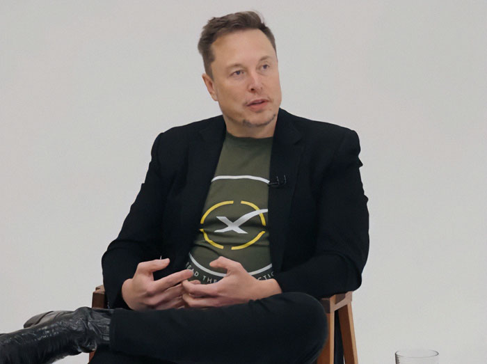 Elon Musk sitting in a black blazer and green SpaceX t-shirt, engaged in conversation during an interview.