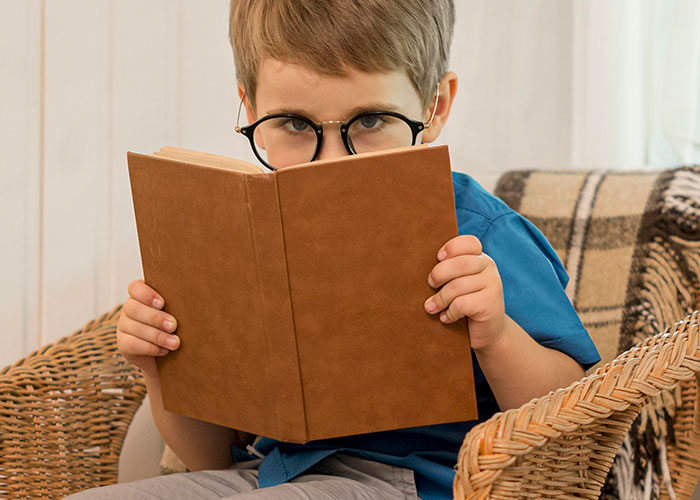 Young boy wearing glasses sitting in a wicker chair reading a brown book about things people did as kids that make them cringe.