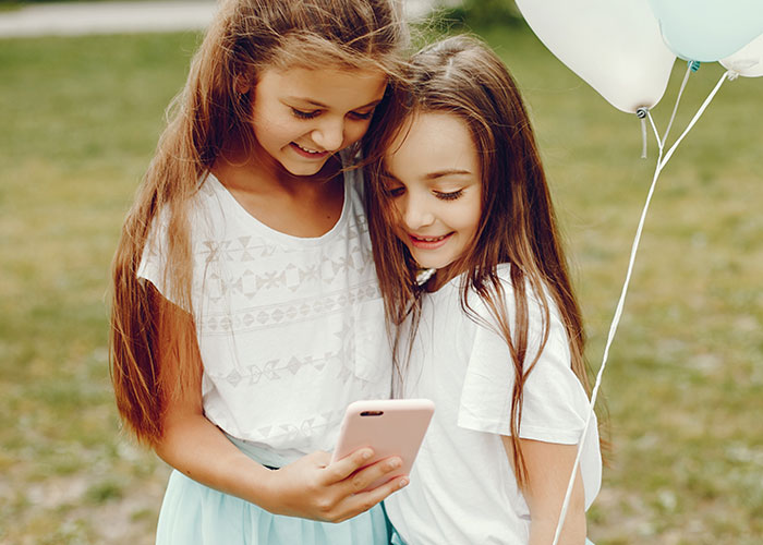 Two young girls smiling and looking at a phone outdoors, evoking childhood memories and playful moments.