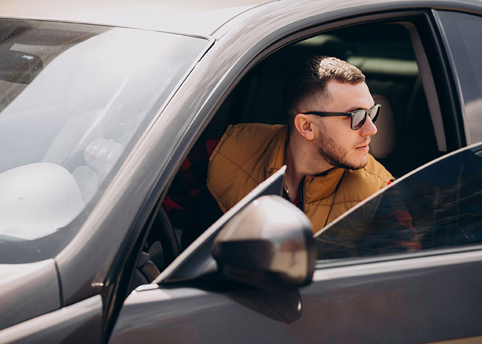 Young man wearing sunglasses and a brown vest leaning out of a car window reflecting on things people did as kids that make them cringe.