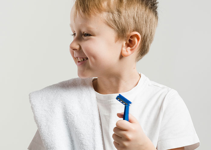 Young boy smiling with a towel over his shoulder, holding a razor, recalling childhood cringe moments.