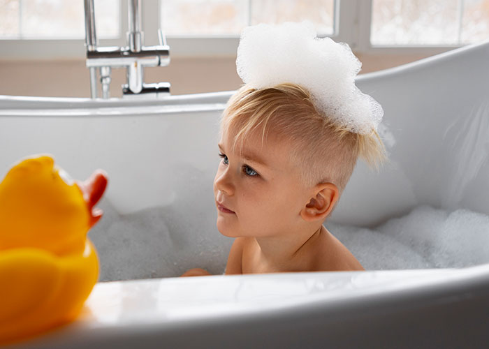 Young child in a bubble bath with foam on head, playing with a yellow rubber duck, recalling childhood cringe moments.