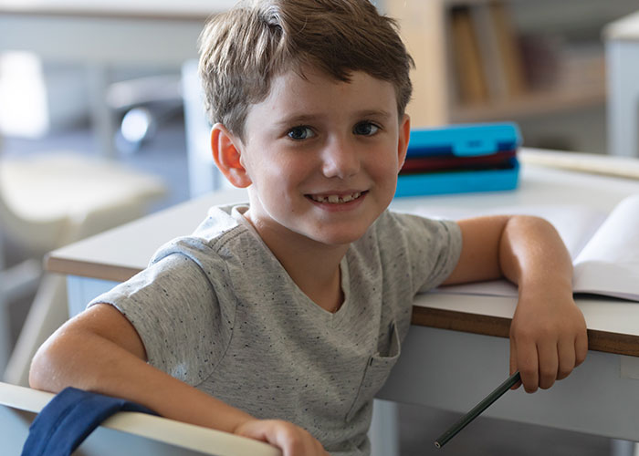 Smiling young boy sitting at desk holding pencil, recalling childhood memories that make him cringe to this day.
