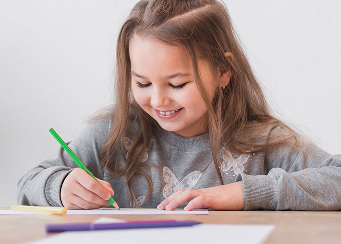 Young girl smiling and drawing with a green pencil, reflecting on childhood things that make people cringe today.