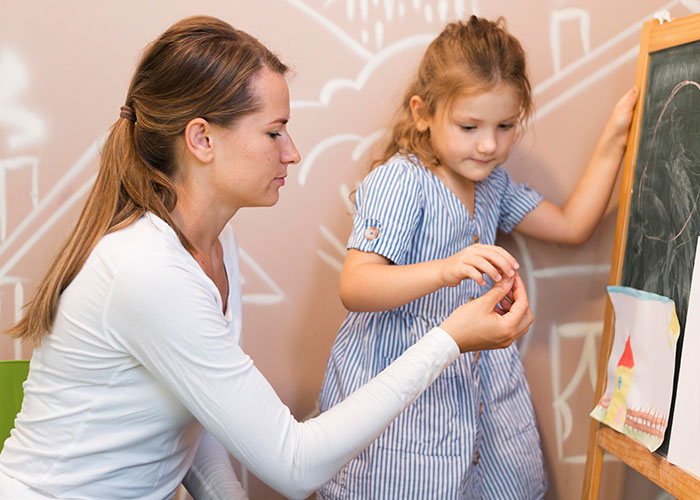 Woman teaching young girl at chalkboard, illustrating childhood moments that make people cringe to this day.