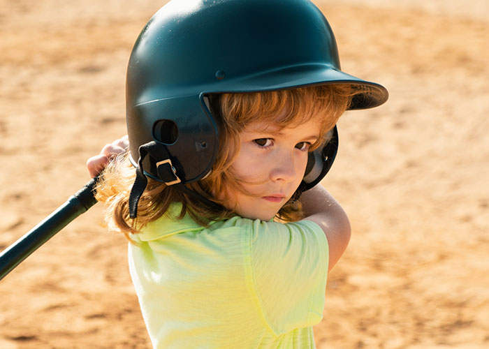 Young child wearing a helmet and holding a bat, representing childhood memories that make people cringe today.