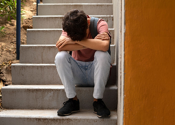 Teen sitting on concrete stairs with head down and arms crossed, reflecting on childhood cringe moments.