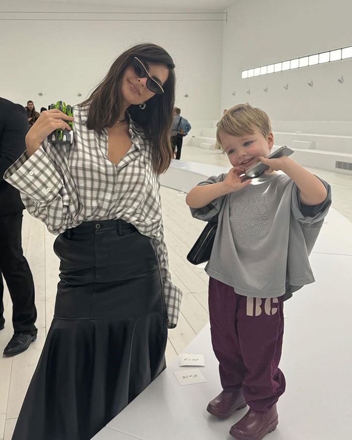 Victoria&rsquo;s Secret model wearing sunglasses and a plaid shirt posing with a smiling child in a modern white space.