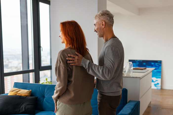 Brother and sister standing inside a house near a window, the brother with a hand on her shoulder, enjoying the ride view.