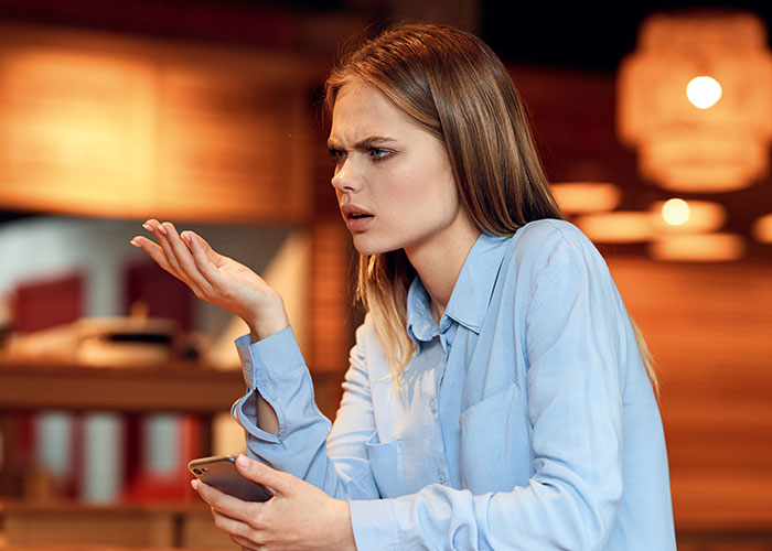 Woman with concerned expression holding phone, confused about inviting some members of friend group for dinner.