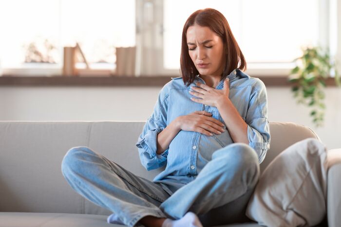 Young woman sitting on a couch clutching her chest and stomach, showing signs of medical problems and discomfort.