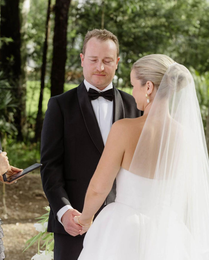 Bride and groom holding hands during outdoor wedding ceremony with bride wearing veil and groom in black tuxedo.