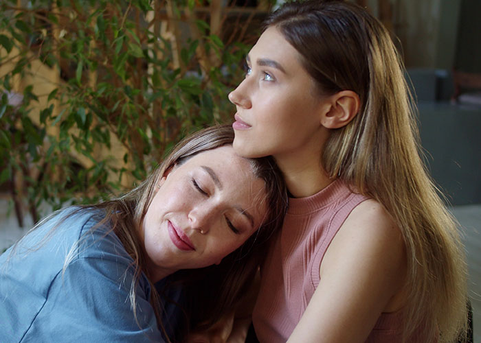 Two women sitting indoors, one resting her head on the other's shoulder, capturing family separate drive Disneyland moment.