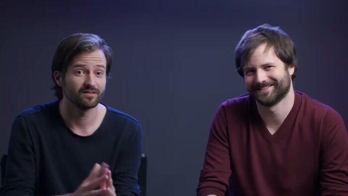 Two celebrity twins sitting against a dark background, one wearing a black shirt and the other in a maroon shirt, smiling.