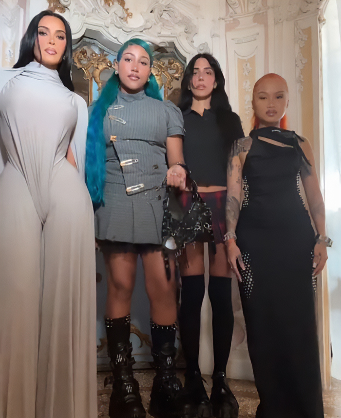 North West with fake face tattoos, piercings, and grills posing with three women in stylish outfits indoors.