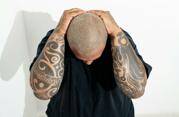Man with shaved head and tattooed arms wearing black shirt holding his head against white background, tattoo artists refused to ink.